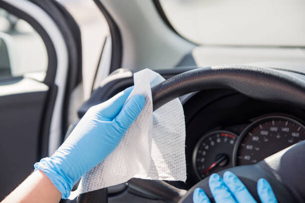 Female hands with blue glove wiping car steering wheel with disinfectant wipe. Horizontal outdoors close-up with copy space.